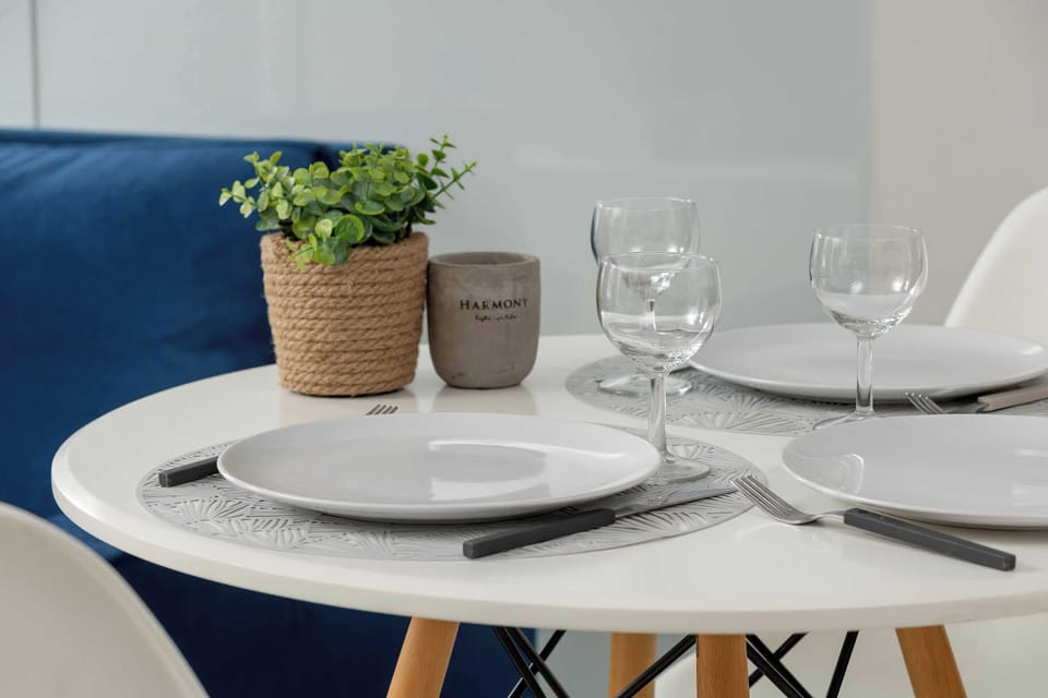 A close-up shot of the dining table shows elegant place settings with gray plates and wine glasses, accented by a small potted plant. The blurred background hints at the comfortable living area nearby.