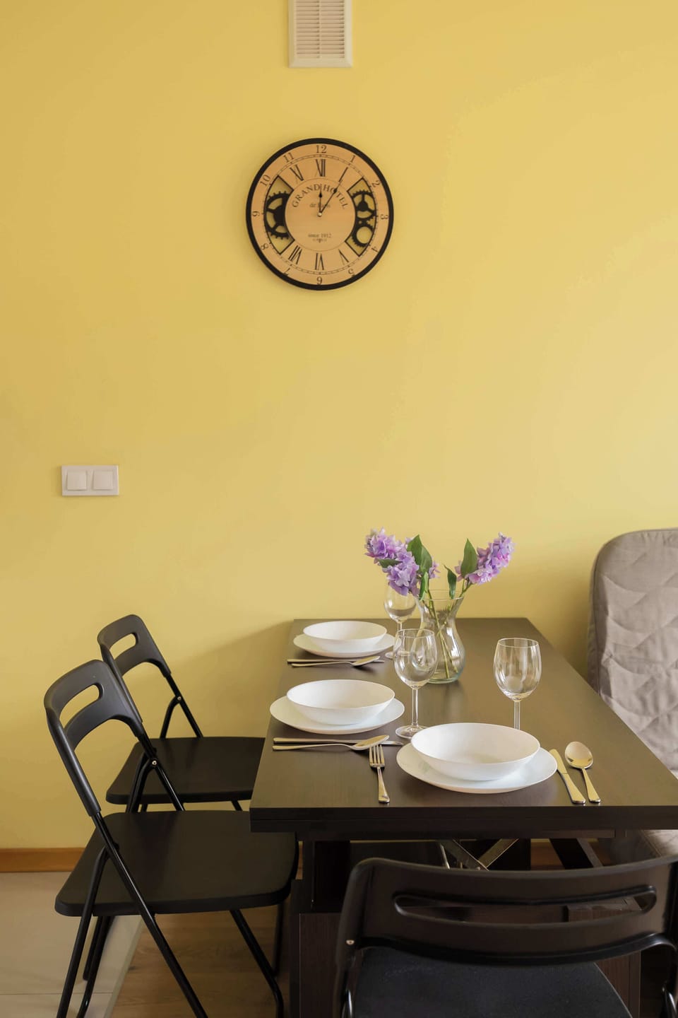 A vertical shot focuses on the dining table, highlighting the warm yellow wall and a circular clock above. The wooden flooring extends throughout the entire living space.
