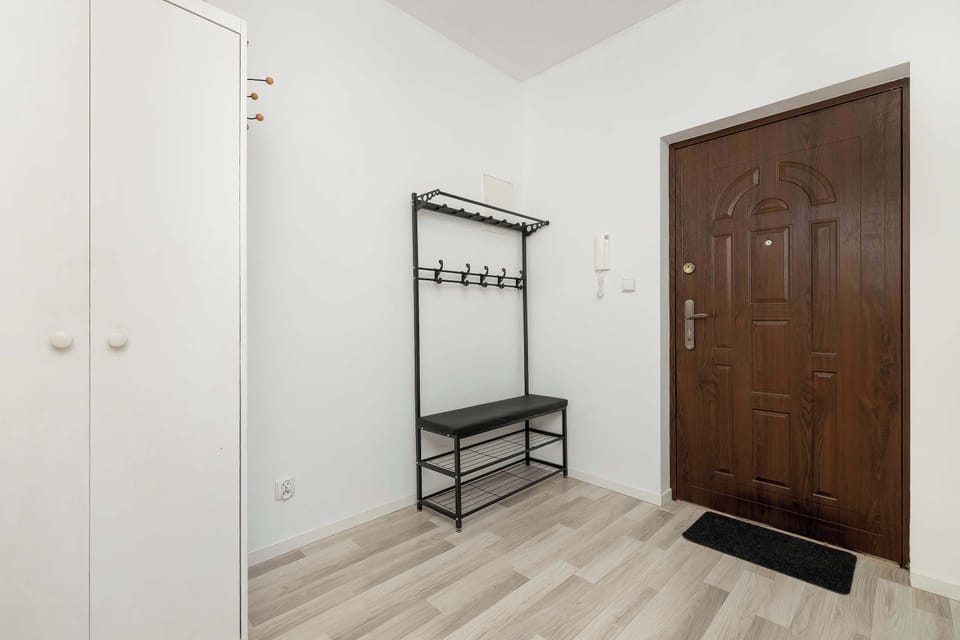 A minimalist hallway features a white wardrobe and a black metal coat rack with a shoe bench. The dark entrance door contrasts sharply with the bright white walls and light floors.