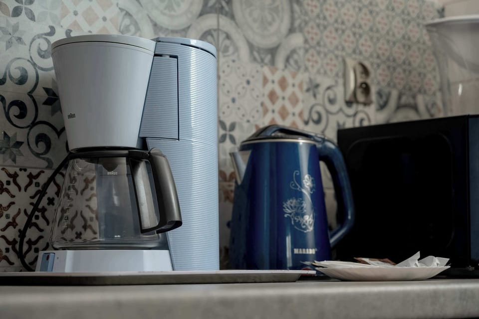 A close-up of a kitchen counter with a coffee maker and an electric kettle placed side by side. The neutral backsplash and compact arrangement emphasize practicality.