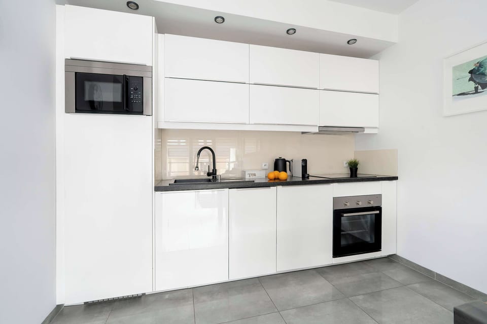 The sleek, all-white kitchen is designed with minimalist cabinetry and dark countertops for a sharp contrast. High-end built-in appliances, including an oven and microwave, maximize the compact space.
