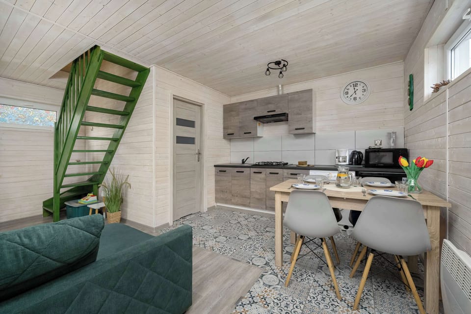 A cozy living area with a green sofa placed next to a small dining table and chairs. Light wooden finishes and a slanted ceiling create a warm, attic-like atmosphere. The space feels bright and functional, ideal for relaxing or casual meals.