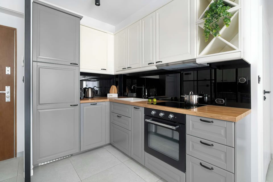 A modern L-shaped kitchenette combines light grey cabinetry with a black tiled backsplash and sleek dark countertops. It is fully equipped with an oven, stovetop, and integrated appliances, maximizing the utility of the corner space.