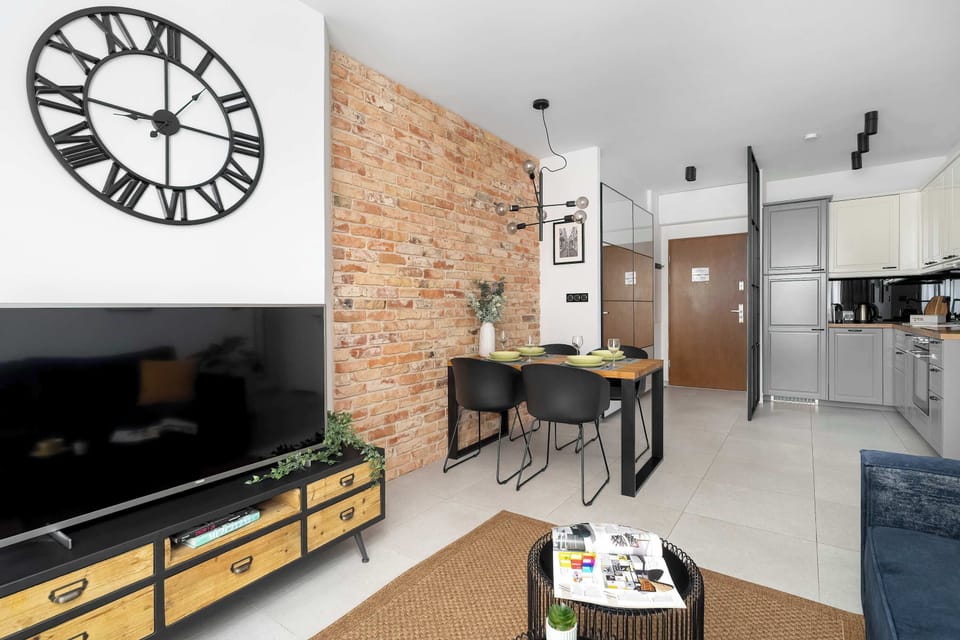 A different angle of the living room showcases the textured brick wall and the TV area, with a view leading towards the entrance and kitchen. The combination of industrial materials and soft textiles creates a balanced interior.