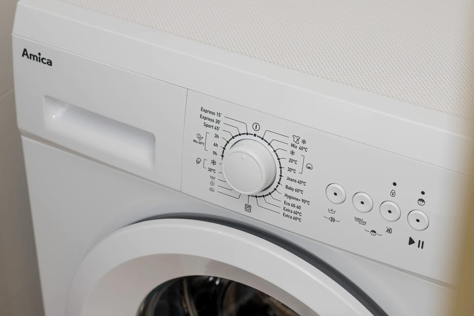 A white front-loading washing machine is tucked into a functional corner of the apartment. Its control panel is clearly visible, suggesting convenient laundry facilities for long-term stays.