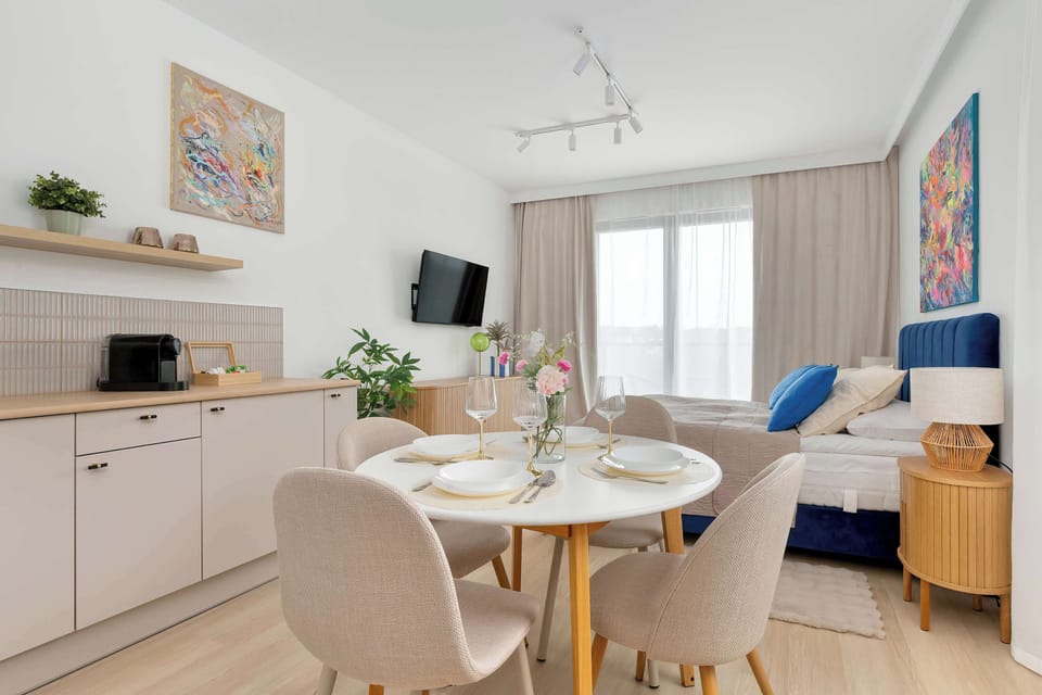 The open-plan living area includes a round dining table with four chairs positioned near a compact kitchenette. Soft natural light flows in through large floor-to-ceiling windows covered by neutral curtains.