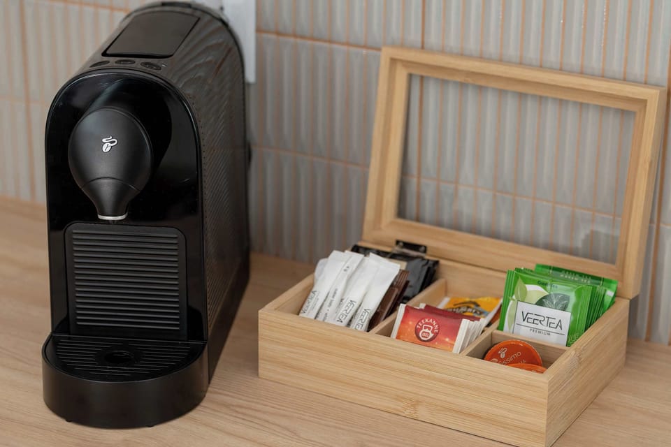 A dedicated coffee station includes a black capsule machine and a wooden organizer box filled with various tea bags. This setup is placed conveniently on the wooden countertop for easy access.