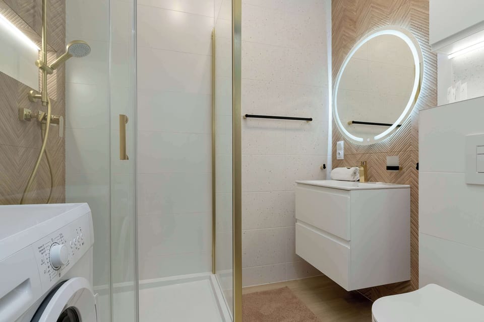 The bathroom features a glass-enclosed shower stall alongside a white vanity unit and a glowing circular mirror. Neutral tiles and warm lighting create a spa-like atmosphere.