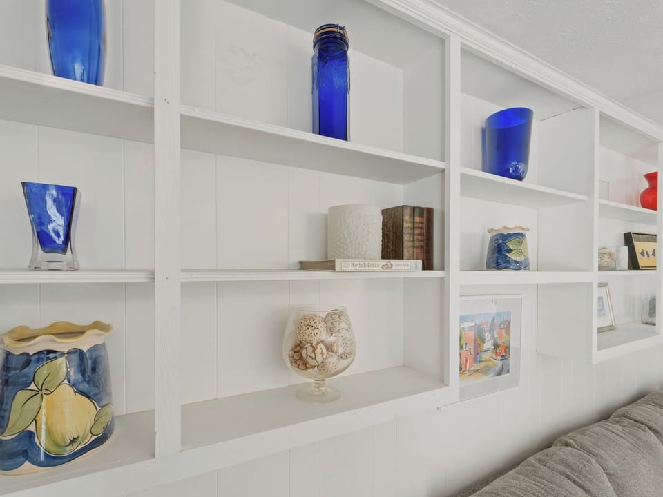 Built-in white shelving displays coastal-inspired decor, from vibrant blue glass pieces to seashell collections, creating an elegant and personalized touch to your living space.