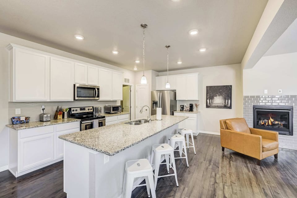 From this angle the kitchen opens toward the living area, with the granite island, white shaker cabinetry, and stainless appliances all visible alongside the fireplace beyond. A space that feels equally suited to cooking and gathering.
