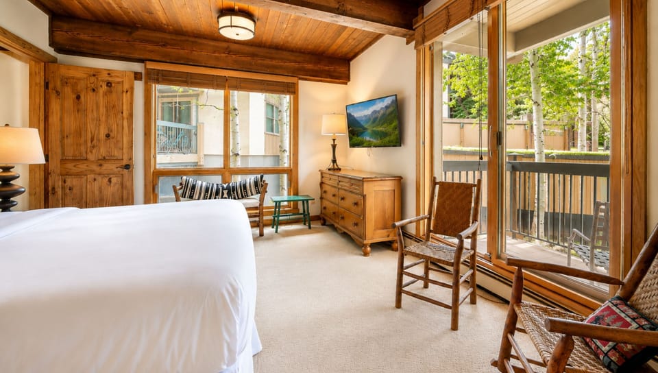 Step into your cozy mountain retreat with vaulted wood ceilings, a plush bed, and your own private balcony framed by tall trees. Unwind in the rocking chair or curl up on the window bench—your peaceful getaway awaits.