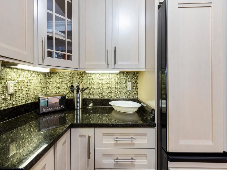 Black granite countertops meet light wood cabinets with glass accents, illuminated by under-cabinet lighting.