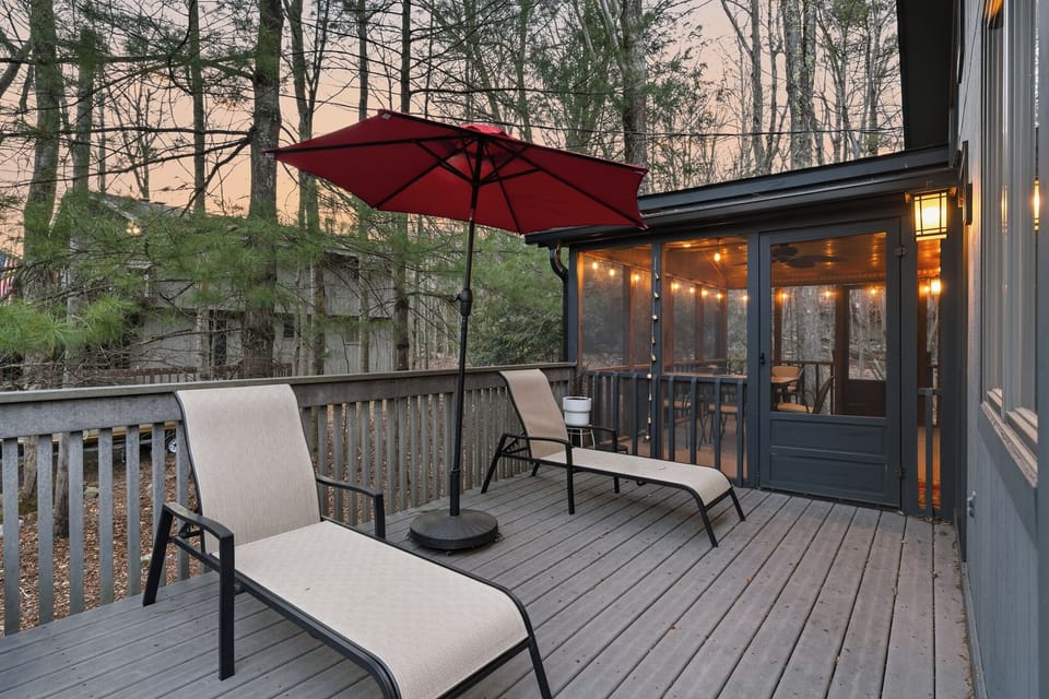 Cozy deck retreat with lounge chairs and a glowing string‑lit porch, surrounded by tall trees at dusk.