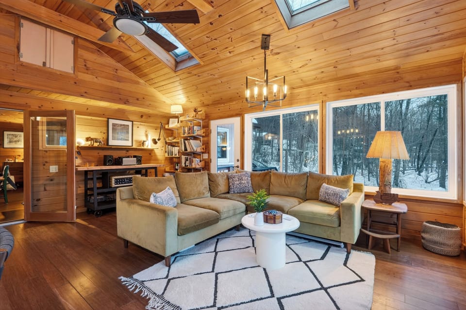 Sun-filled living area with wall-to-wall windows, vaulted wood ceilings, and forest views—an airy, comfortable space to relax and reconnect indoors.
