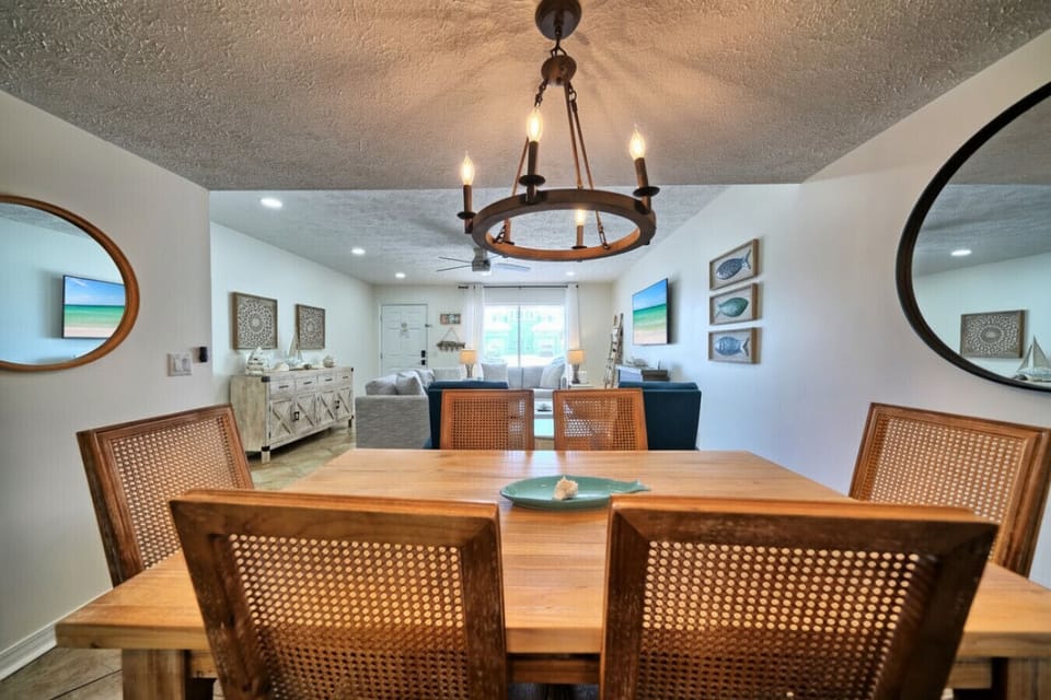 This stylish dining area features a rustic wooden table with seating for four, illuminated by a charming chandelier. It offers a clear view into the bright, open living space, showcasing coastal decor and a flat-screen TV.