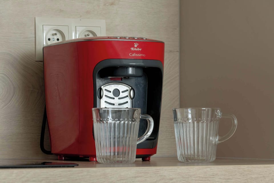 A close-up view features a red Nespresso coffee machine with two glass mugs ready for use. A framed wall outlet is also visible. The simple setting suggests a quick and easy coffee station.