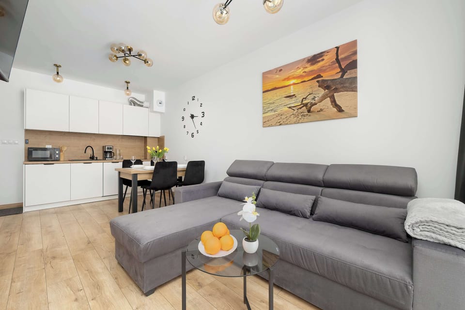 This wide-angle shot shows the seamless transition between the grey sofa and the kitchen area in the open living space. A glass coffee table holds a bowl of oranges, adding a splash of color to the neutral palette.