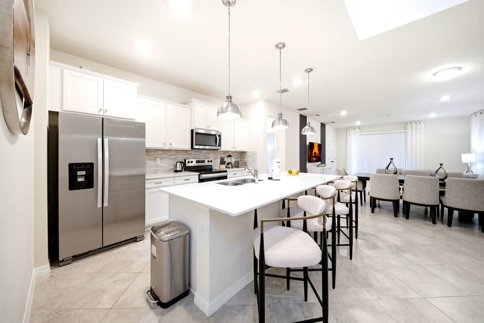 This bright kitchen boasts stainless steel appliances, a spacious island with stylish seating, and elegant light fixtures, all creating a warm atmosphere with neutral tones and polished tile flooring, ideal for your culinary.