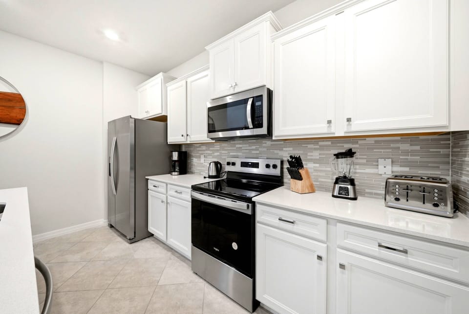 This bright kitchen features sleek white cabinetry and modern stainless steel appliances for a functional meal prep space. The elegant gray backsplash adds sophistication, making it a delightful spot to cook and socialize with.