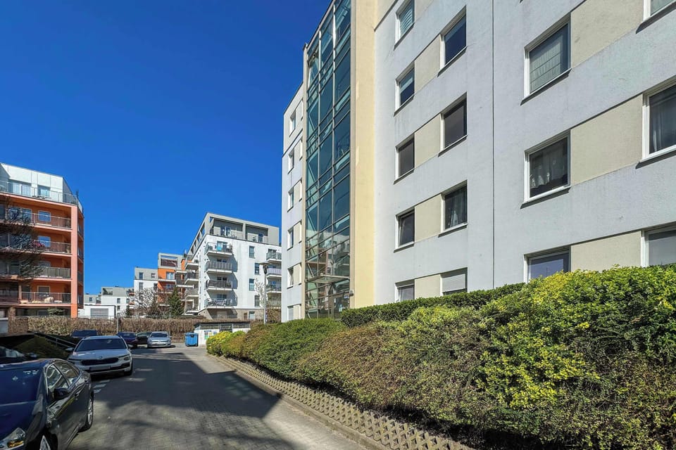 The exterior facade of the apartment building is shown, featuring clean white walls and multiple glass-paneled balconies. A row of neatly trimmed green hedges lines the base of the structure.