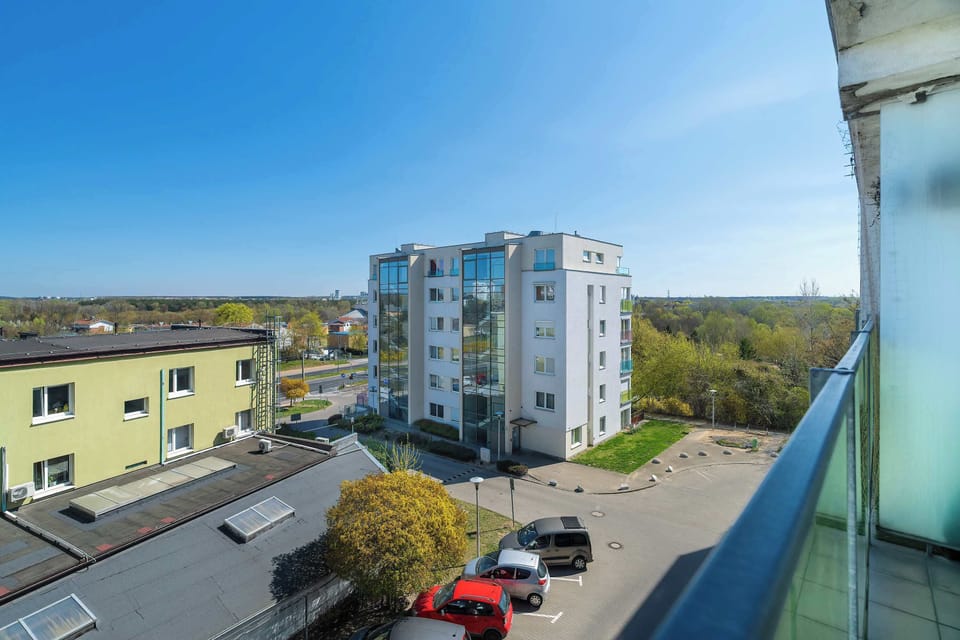 An elevated view from the balcony captures the surrounding residential buildings and green treetops under a clear sky. The perspective shows the quiet, organized nature of the local street.
