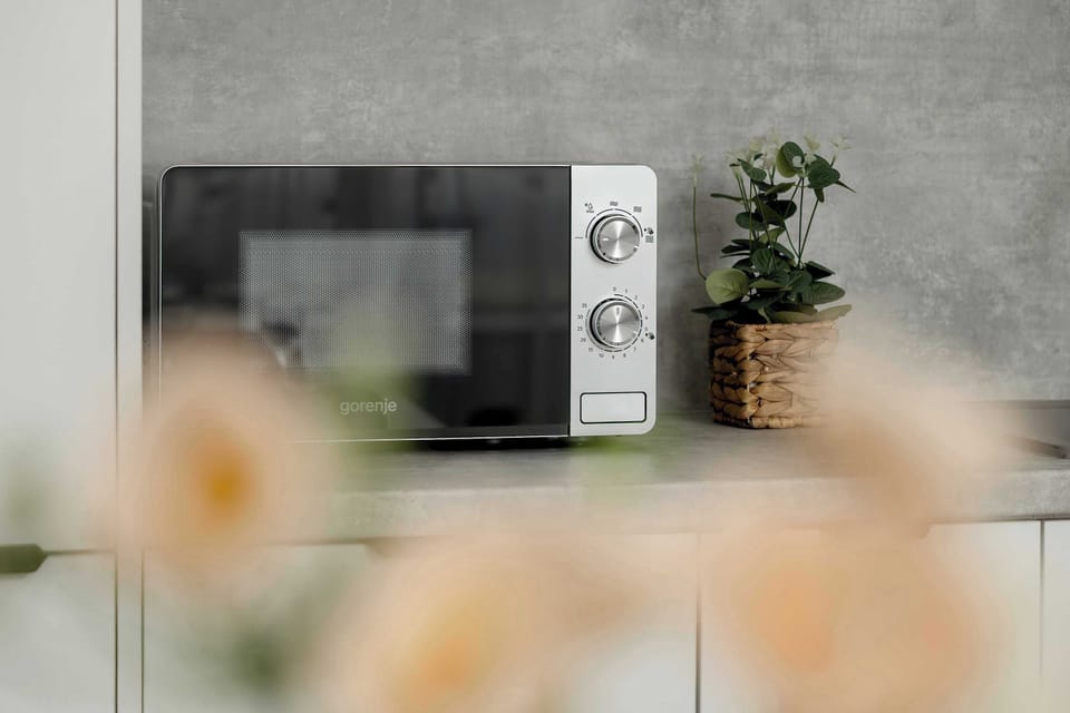A close-up artistic shot shows a black microwave and a small potted plant on the kitchen counter. Blurred floral elements in the foreground add a soft, lifestyle-oriented feel to the image.
