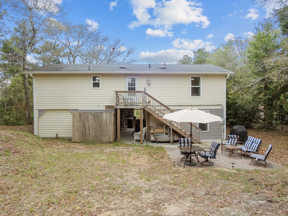 A two-story vacation home featuring a spacious backyard with outdoor seating, umbrella shade, and BBQ grill surrounded by mature trees.