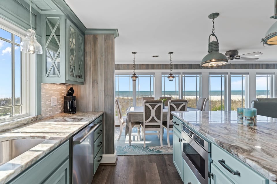 Coastal kitchen with blue cabinets, granite counters, island, stainless appliances, pendant lights, dining area, wide windows, dune views, and ocean scenery.
