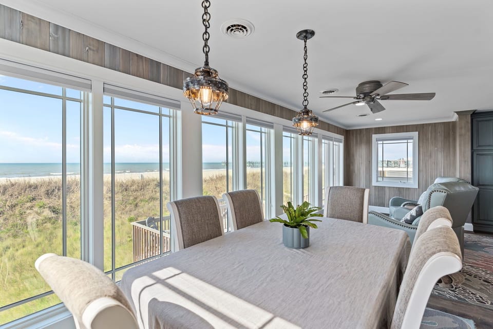 Beachfront dining area with cushioned chairs, pendant lights, wood accent walls, wide windows, dune views, ocean waves, pier view, and nearby sitting space.