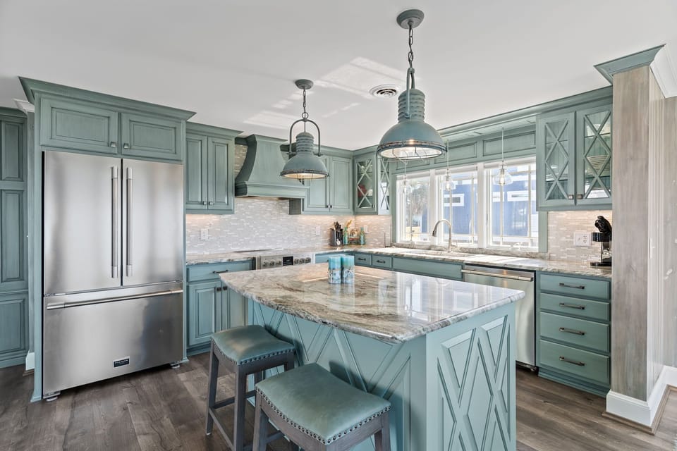 Coastal kitchen with blue cabinets, granite counters, sink, dishwasher, glass pendants, large windows, dining area, wood accents, and ocean views.