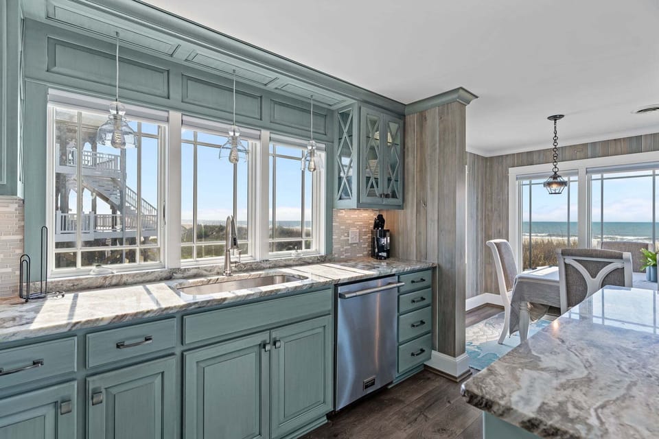 Coastal kitchen with blue cabinets, granite counters, sink, dishwasher, glass pendants, large windows, dining area, wood accents, and ocean views.
