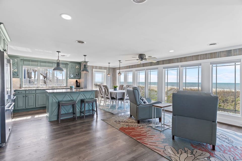 Open beachfront great room with blue kitchen island, bar stools, dining table, leather chairs, pendant lights, wide windows, wood floors, dune views, and ocean scenery.