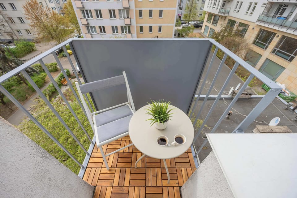 A private balcony offers a charming spot for coffee with a small wooden table and two chairs. The floor is covered in wooden tiles, and a grey privacy screen surrounds the railing.