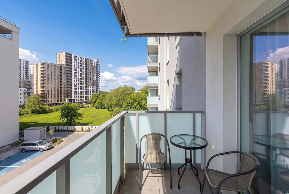A view from a high-rise balcony, complete with a small glass-topped table and two metal chairs, looking out over a city landscape and trees.