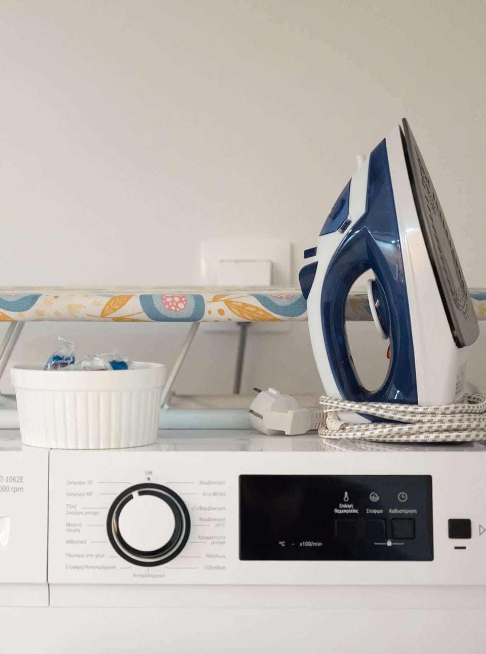 Laundry area with a washing machine, iron, and ironing board neatly arranged. Practical setup offering everything needed for basic laundry and garment care during your stay.