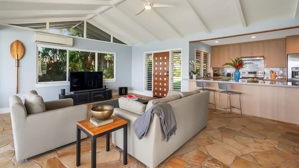 Relax in your open-plan living space with modern kitchen, comfortable seating, and tropical garden views through expansive windows.