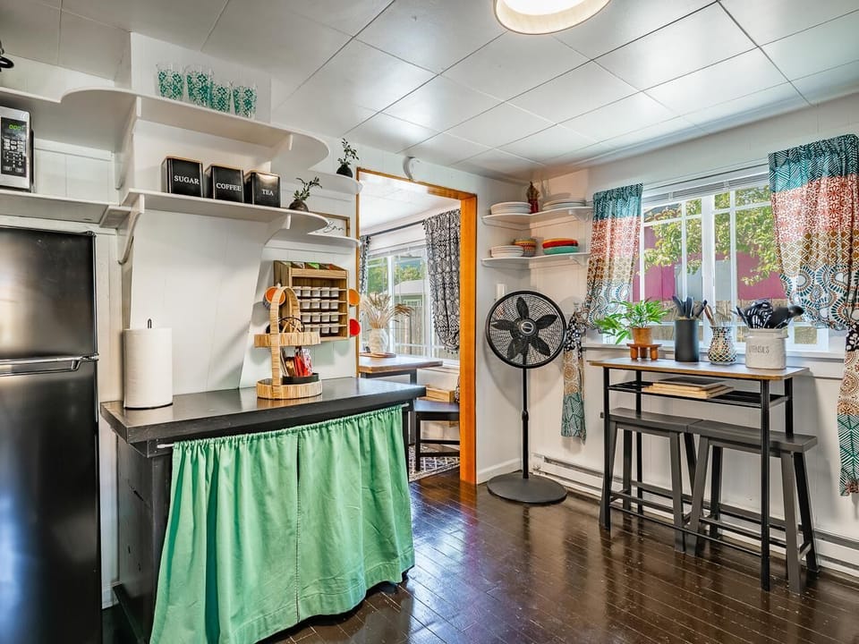 Kitchen corner with black appliances, open white shelves, patterned curtains, and a tall dining table with bench seating.