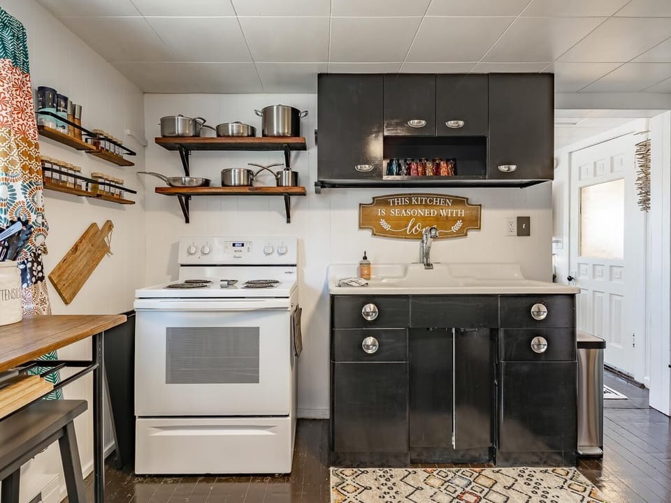 Kitchen with black cabinets, white stove, open wooden shelves holding pots, and a patterned rug on dark floors.