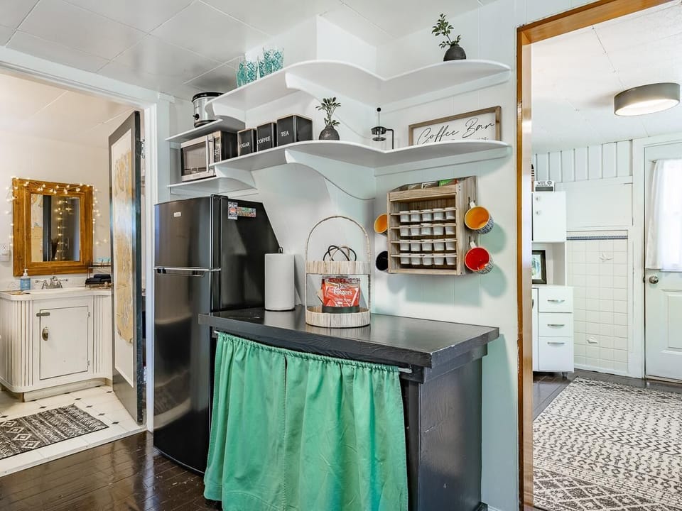 Compact coffee bar with white scalloped shelves, a black refrigerator, and a black counter skirted in green fabric.