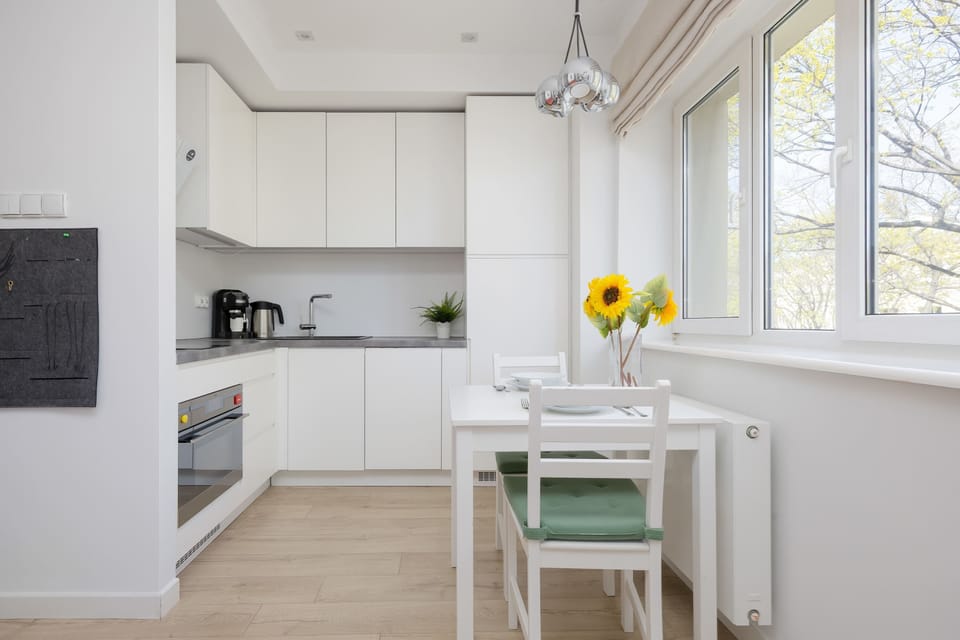 A bright kitchen corner shows the dining table tucked against a radiator and a large window. Green cushions on the white chairs provide a subtle splash of color to the room.