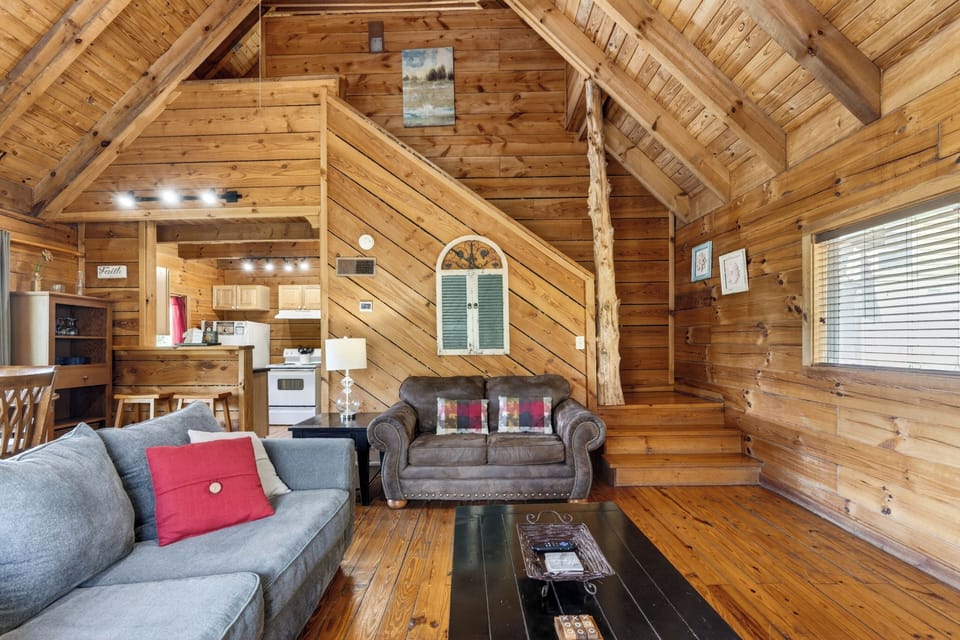 Warm, wood‑lined cabin living room with a stone fireplace, a queen sleeper sofa, soft natural light, and mountain views framed by the dining area windows.