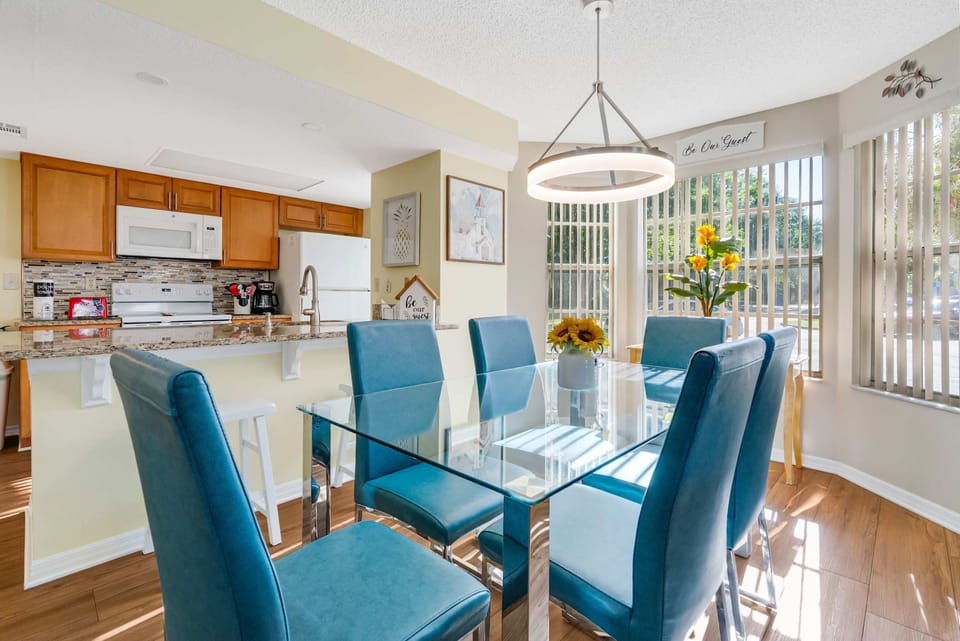 Bright dining area with blue chairs and a glass table next to a modern kitchen.