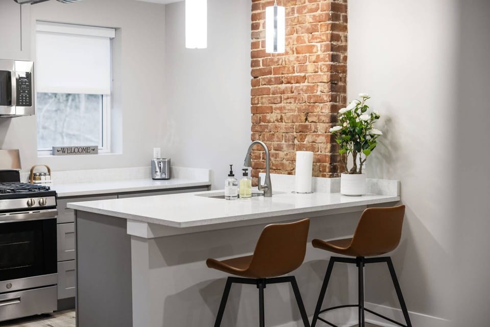 Kitchen island close-up with quartz countertop, exposed brick column, glowing pendant lights, and leather bar stools.