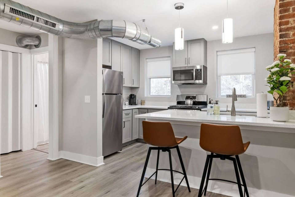 Full kitchen view showing stainless refrigerator, gray shaker cabinets, island seating, exposed ductwork, and brick.