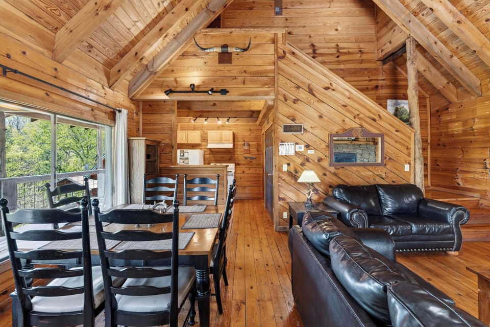 Open‑concept log cabin with warm wood interiors, a cozy dining nook, leather sofas, and sunlight pouring in through the big windows.
