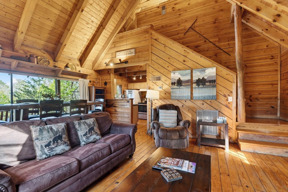 Warm cabin living room with a glowing stone fireplace, a queen sleeper sofa, and sunlight pouring through big mountain‑view windows.