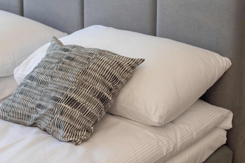 A close-up shot focusing on the bedding shows the edge of a bed with a white-patterned, textured pillow resting against larger white pillows. The grey quilted headboard is visible behind them.