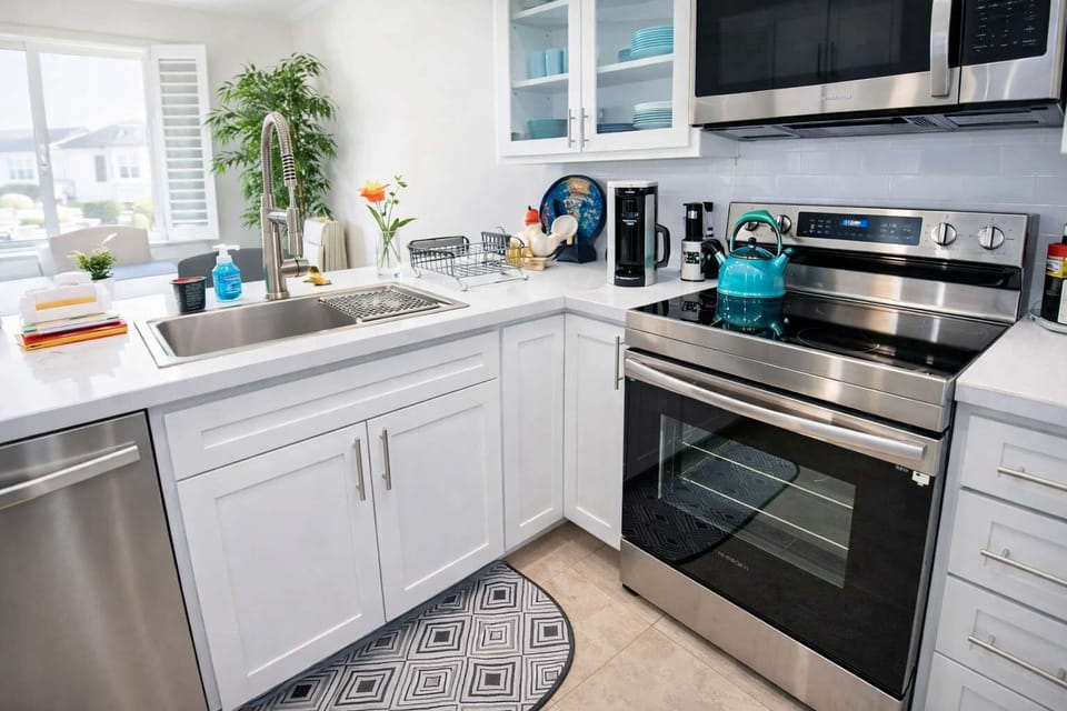 Bright, coastal-inspired kitchen featuring sleek white cabinetry, stainless steel appliances & a turquoise kettle accent. Natural light fills the space, highlighting the clean countertops, fresh florals & patterned rug—perfect for seaside charm.