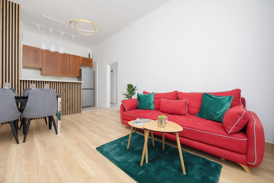 A modern living space featuring a red sofa, a TV unit, and a small dining table nearby. The open layout connects the relaxation and dining zones seamlessly, with light colors enhancing the sense of space.