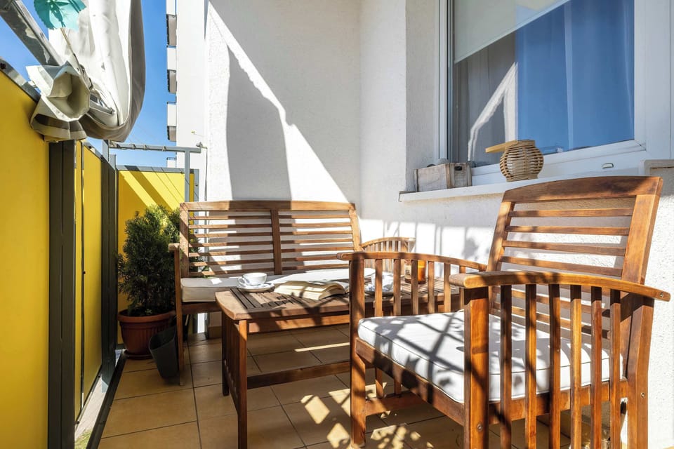 A narrow balcony with wooden chairs and a table, enclosed by yellow walls and open to daylight.